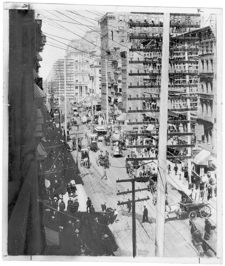 Historical black-and-white photograph of a busy 1890s city street crowded with horse-drawn carriages, pedestrians, and a dense network of overhead telephone and electrical wires crisscrossing between tall poles and buildings.
