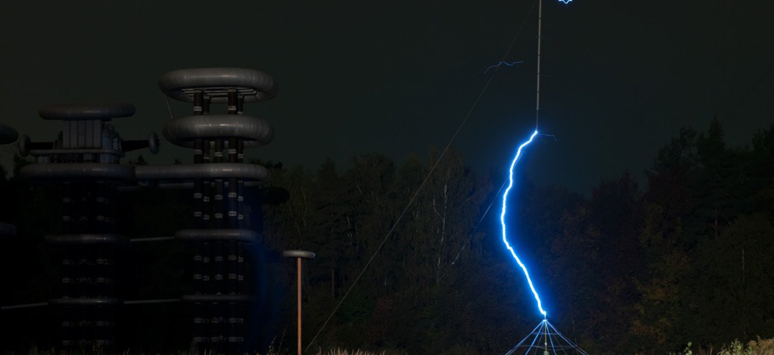 A multi-stage Marx generator firing at night against a forest, producing a bright blue lightning-bolt discharge approximately 100 meters tall.