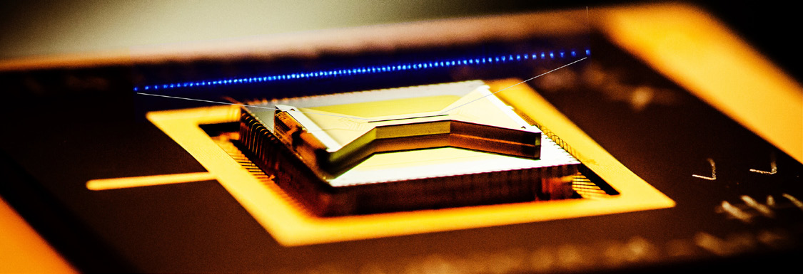 A close-up photograph of a surface ion trap in a gold package with a row of fluorescing trapped ions glowing blue above the chip.