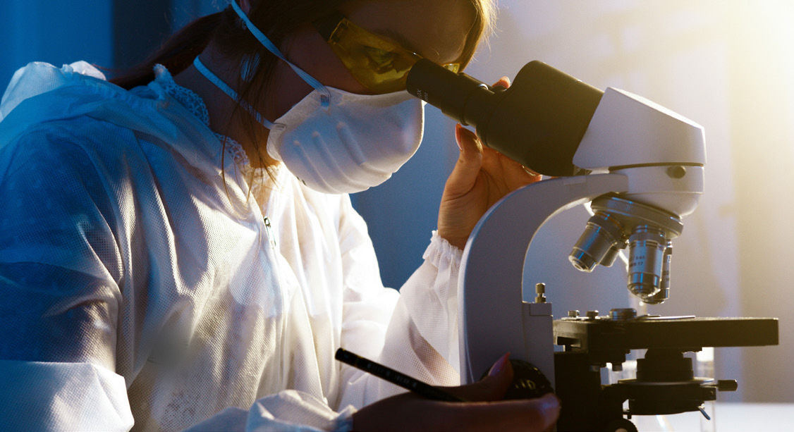 Researcher in protective gear looking through a microscope